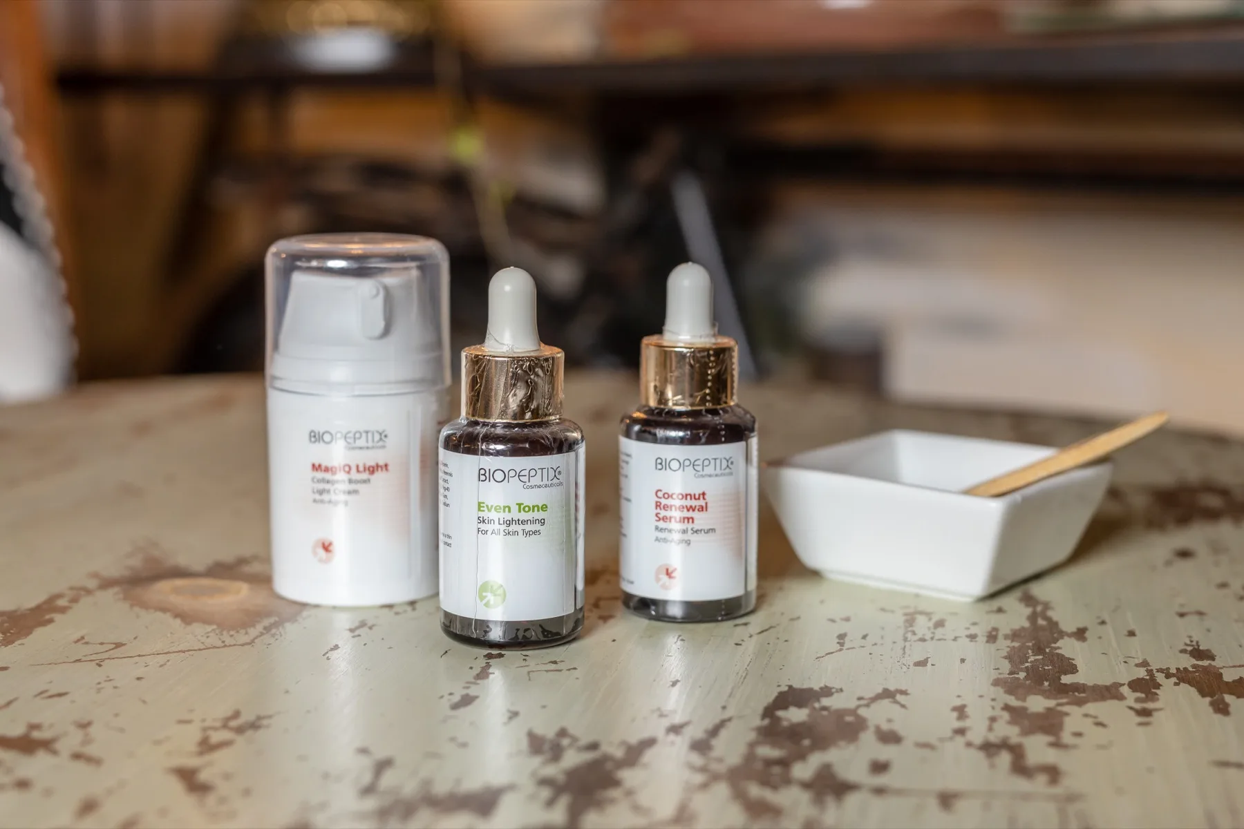 Two amber glass dropper serums and a white pump bottle grouped beside a small porcelain dish on a weathered pale-green vintage table at ESSpa Kozmetika