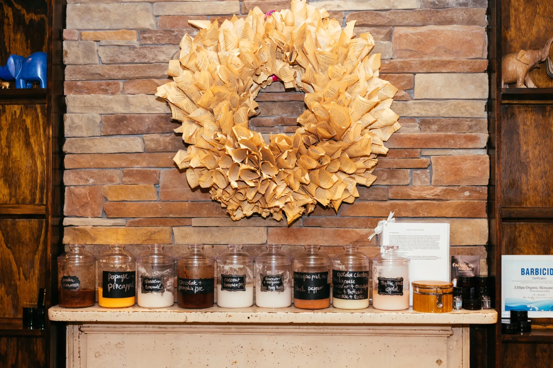 A row of labeled apothecary jars and a lit candle along a wooden mantel beneath a soft ruffled wreath on the stone wall at ESSpa Kozmetika