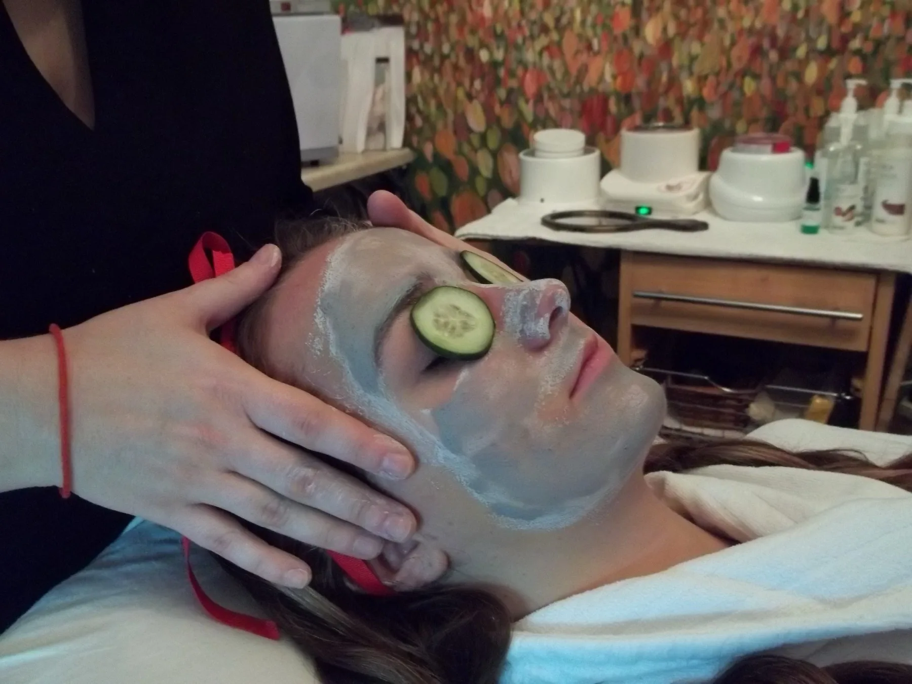 Therapist's hands cradling a guest's face during a cleansing mask facial at ESSpa, a cucumber slice resting over one eye, treatment products lined up on the shelves behind