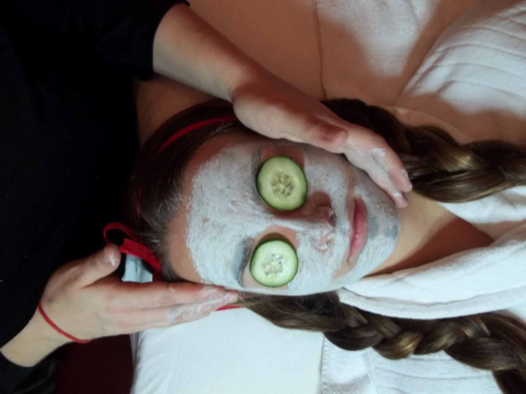 Guest resting under a full facial mask with cucumber slices over both eyes while the esthetician's hands rest gently at the forehead and cheek