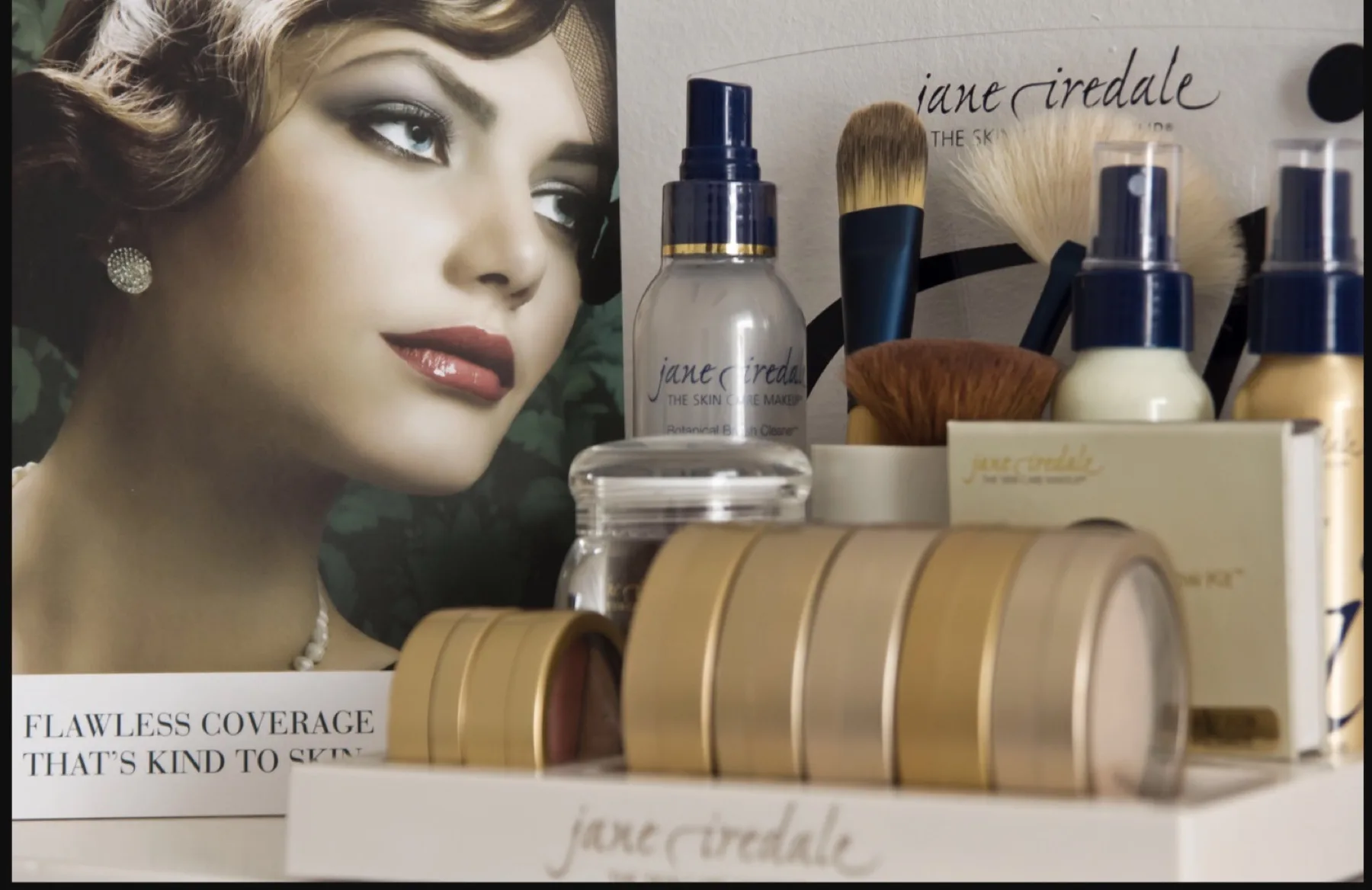 Jane Iredale mineral makeup display at ESSpa Kozmetika — gold compacts, brushes and setting sprays arranged beside a vintage-style beauty poster
