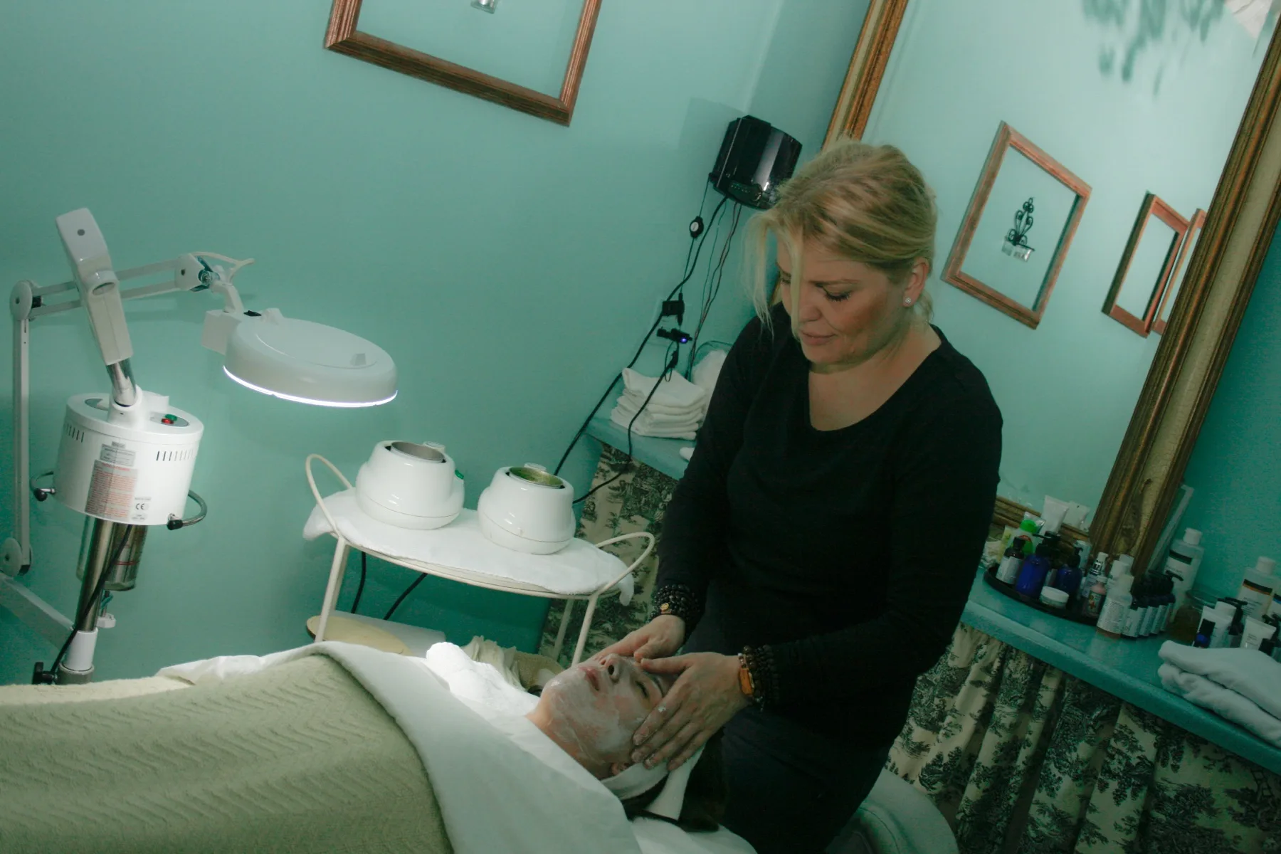 ESSpa esthetician performing a hands-on anti-aging facial on a client resting under a soft green blanket in the calm mint-toned treatment room, magnifying lamp and steamer at the ready
