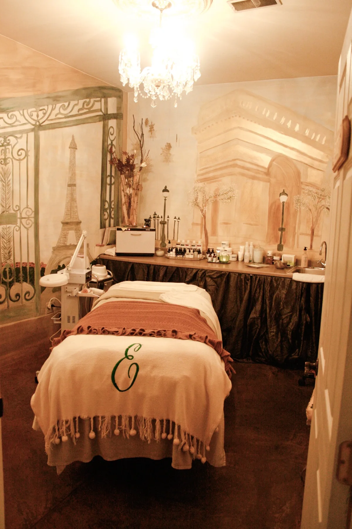 A treatment table dressed in soft linens and a monogrammed throw, waiting under the chandelier in an ESSpa treatment room