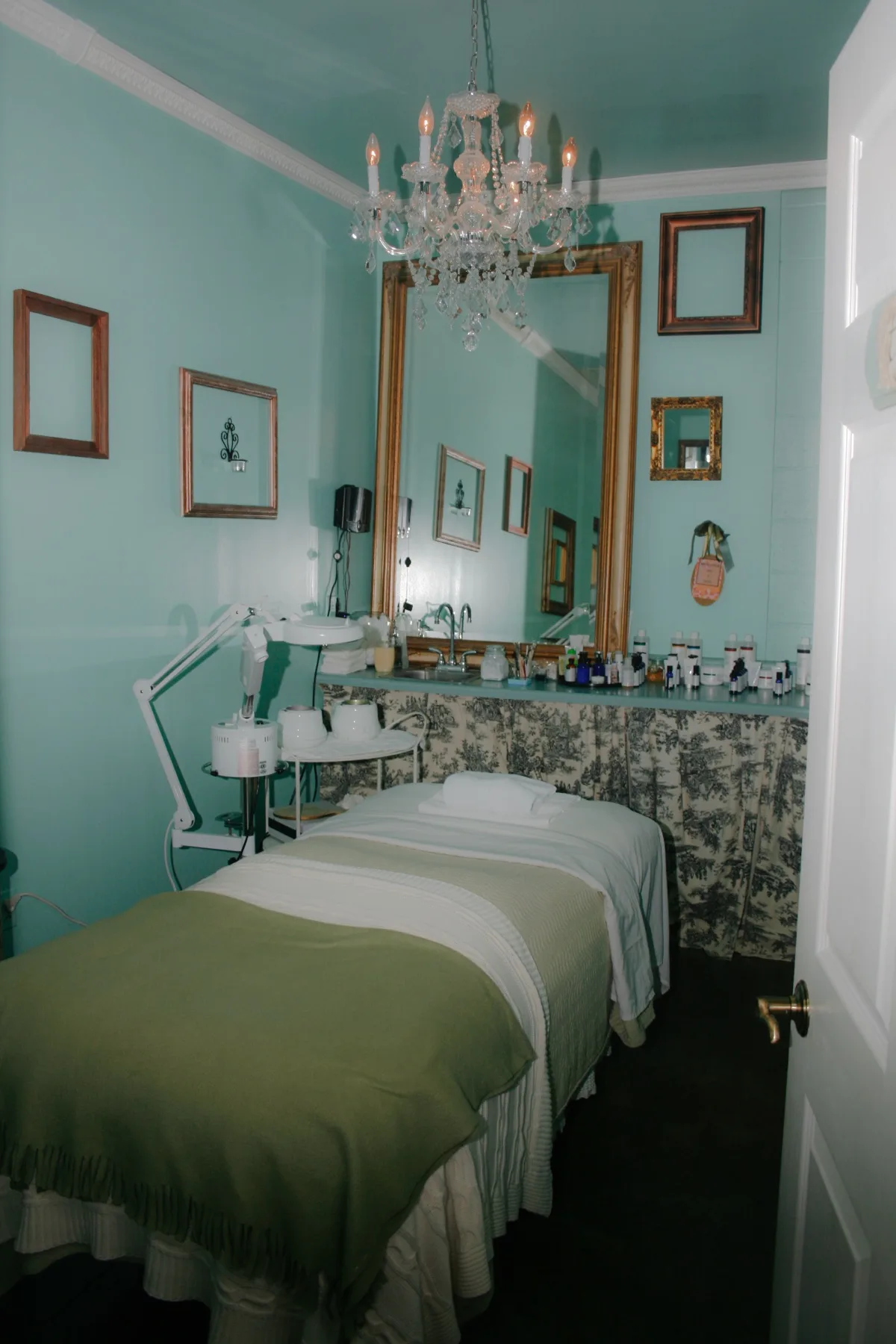 A serene mint-green treatment room at ESSpa in Aspinwall, with a draped treatment table and an adjustable therapy lamp beneath a crystal chandelier