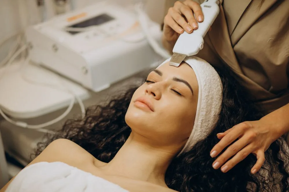 Esthetician gliding a slim resurfacing wand across a relaxed guest's face in our treatment room, with the facial unit at her side